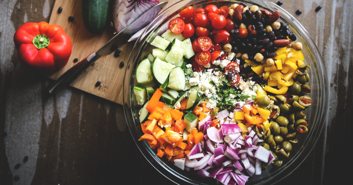 Colorful Mediterranean diet salad in a glass bowl with chopped tomatoes, beans, chopped cucumbers, chopped peppers, halved olives, chopped red onions, and feta cheese crumbles.