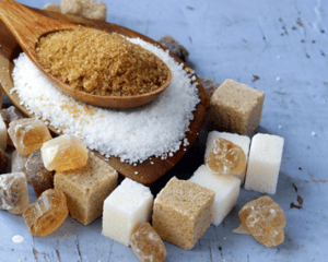 Various types of sugar in wooden spoons and in cube and rock form on a rustic white table.