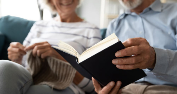 Senior female knitting seated beside a senior male reading a book.