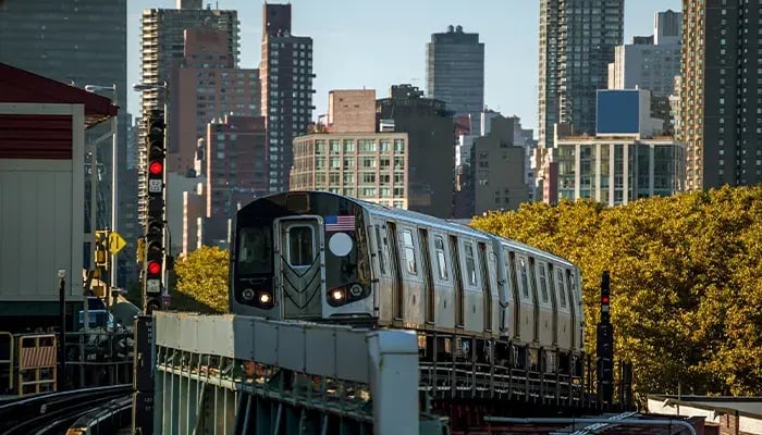 New York City subway train on an elevated track, with skyscrapers and autumn-colored trees in the background.