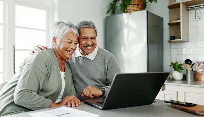 Smiling senior couple sitting together in a bright kitchen, looking at a laptop screen while engaging in a virtual conversation